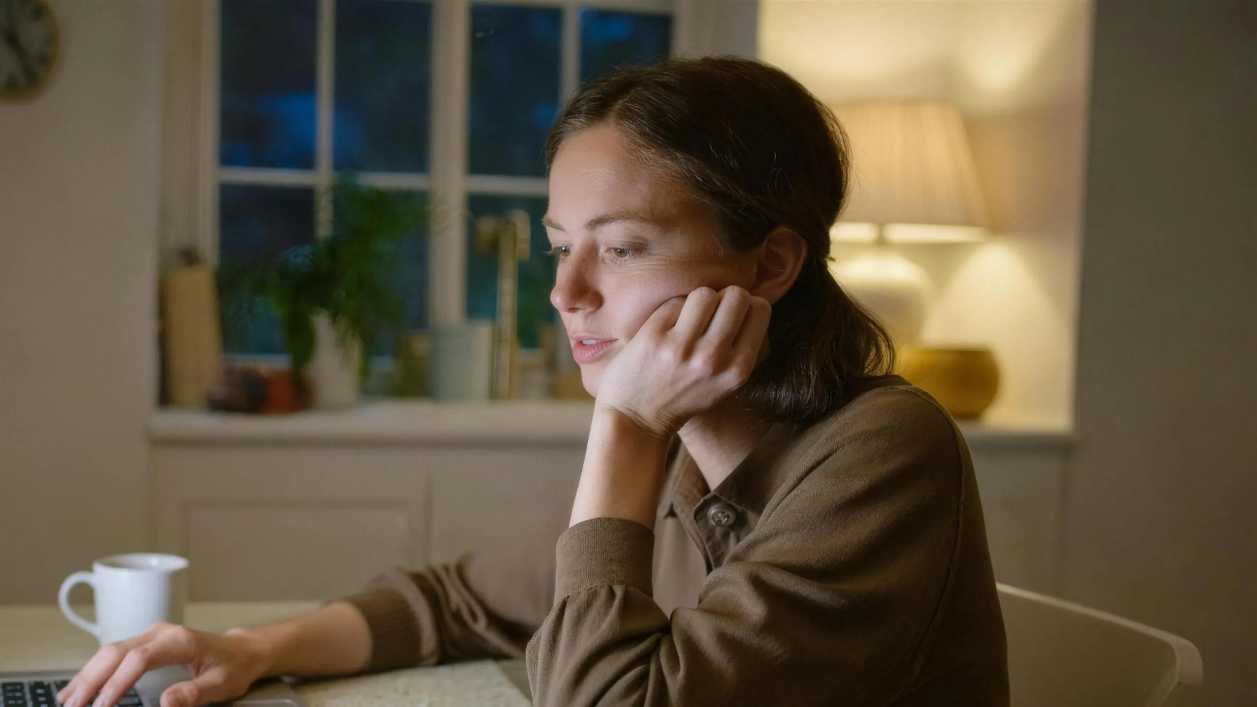 A woman at her kitchen table in the evening, laptop open, hesitating.