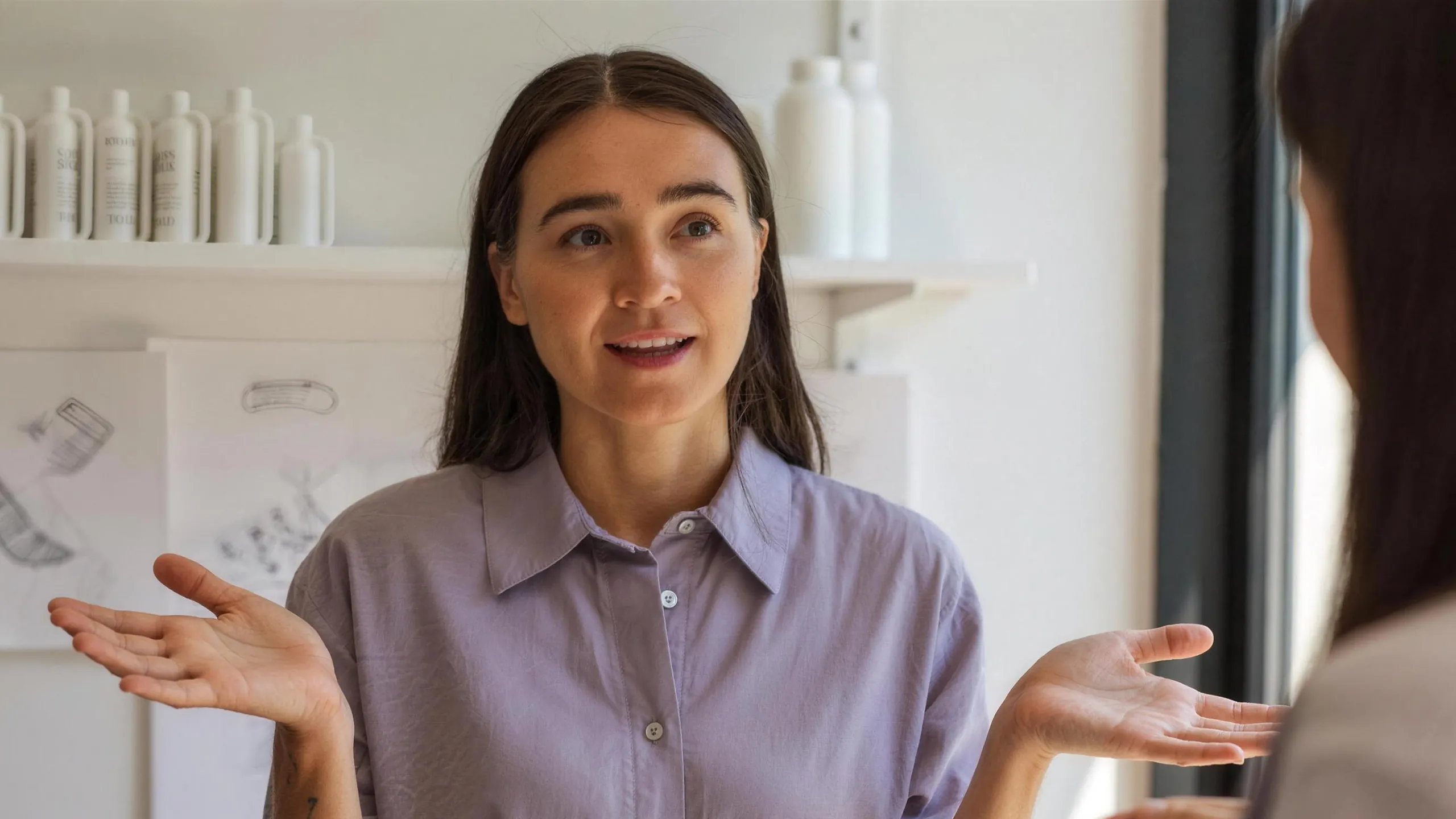 A customer in a product design studio, describing with her hands the product she wishes existed, sketches and prototypes blurred behind her.