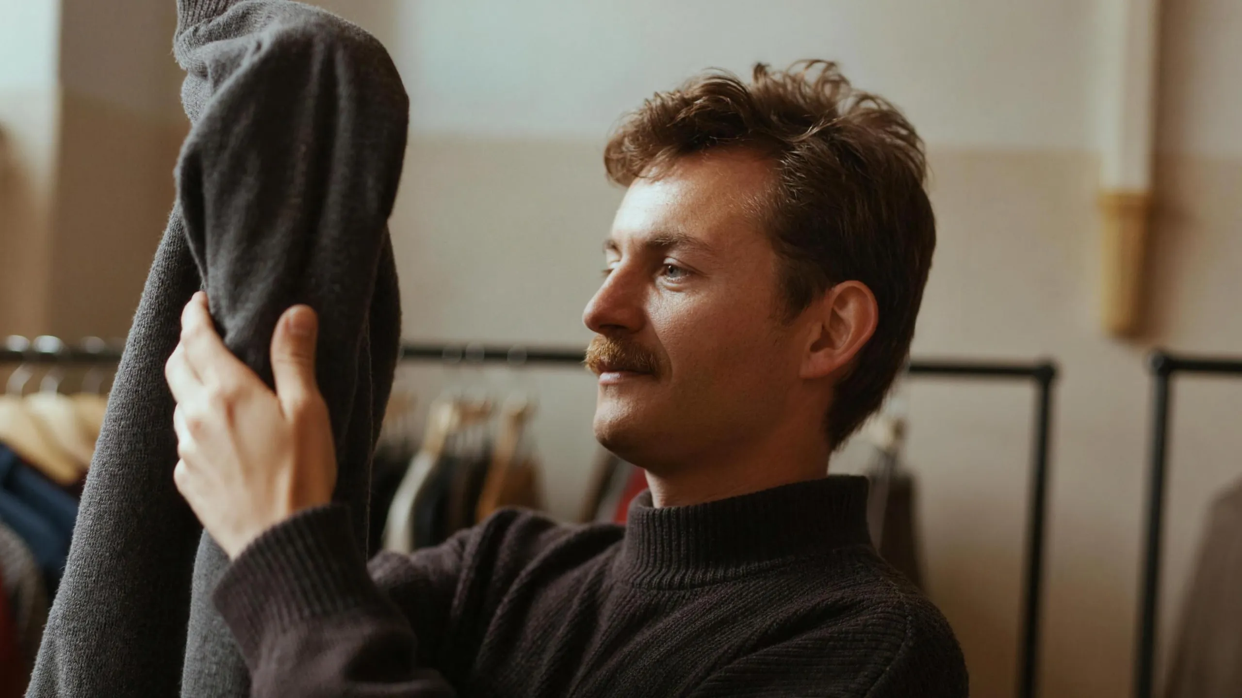 A man in a clothing shop feeling the sleeve of a plain wool jumper, judging whether it is worth the price.